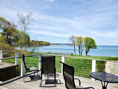 A serene view of a lake from a deck, featuring green trees and a clear blue sky.