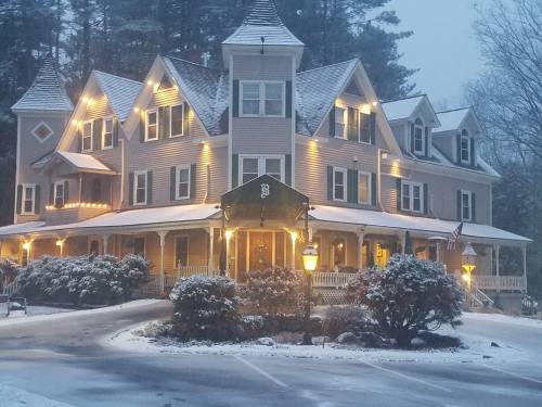A charming, snow-covered Victorian house illuminated with warm lights, surrounded by trees in a winter landscape.