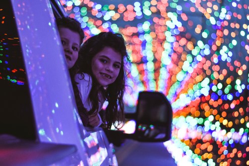 Two children peek out of a car window, surrounded by vibrant, colorful holiday lights.