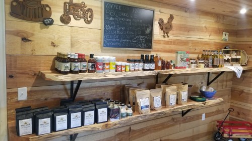 A wooden shelf displays various jars, bottles, and bags of local products in a rustic shop with a chalkboard menu.