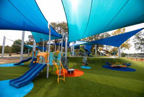 Colorful playground with slides and climbing structures, shaded by blue canopies, set on green grass.