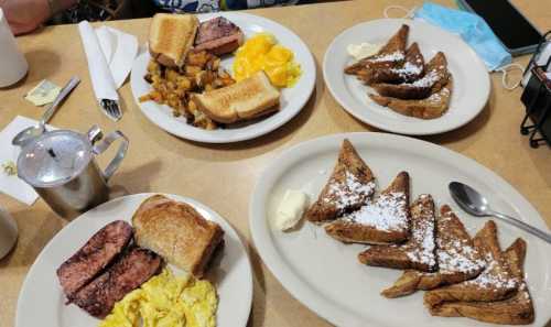 A table with two plates of breakfast: one with eggs, ham, toast, and potatoes; the other with French toast and butter.