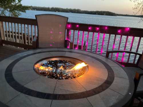 A fire pit with flames sits on a deck overlooking a lake, illuminated by pink lights at dusk.