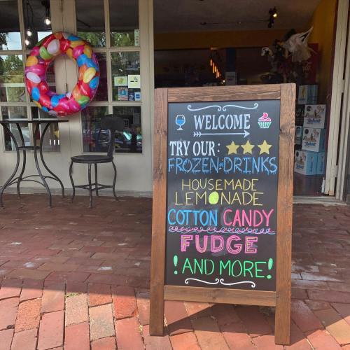 A chalkboard sign outside a shop welcomes visitors and advertises frozen drinks, lemonade, cotton candy, and fudge.