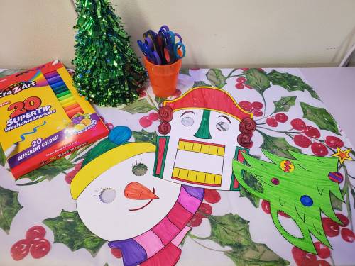 Colorful holiday-themed paper masks of a snowman, nutcracker, and Christmas tree on a festive tablecloth with art supplies.