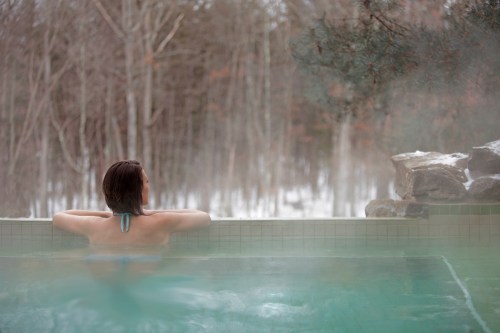 A person relaxes in a steaming hot tub surrounded by a snowy forest landscape.