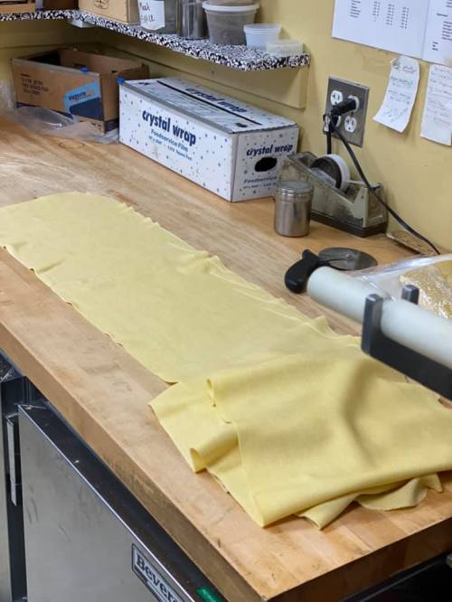 A wooden countertop with a long sheet of fresh pasta and kitchen supplies in the background.