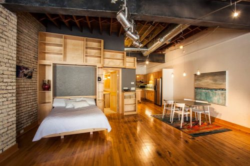 Cozy studio apartment with a bed, dining area, and modern kitchen, featuring wooden floors and exposed brick walls.