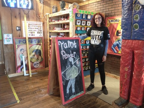 A person stands next to a chalkboard sign that reads "Tarot Reader" in a colorful, eclectic space.
