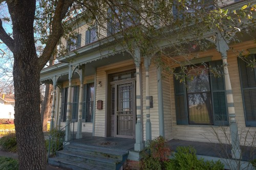 A historic house with a porch, decorative trim, and large windows, surrounded by trees and greenery.