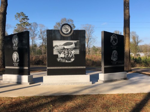 A memorial with three black granite panels featuring engraved images and text, surrounded by trees and blue sky.