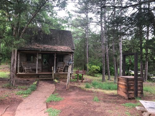 A rustic wooden cabin surrounded by trees, with a stone path leading to the entrance and a wooden sign nearby.