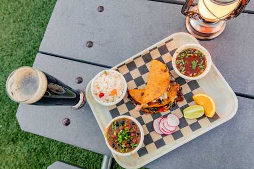 A wooden tray with tacos, rice, beans, and salsa, accompanied by a dark beer and a lantern on a picnic table.