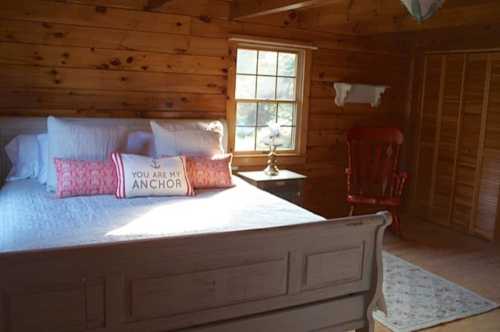 Cozy wooden bedroom with a bed, decorative pillows, a lamp, and a red rocking chair by the window.