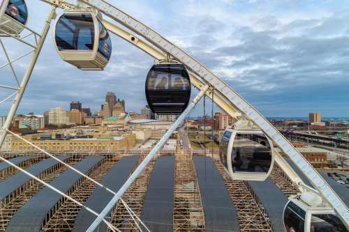 A view of a Ferris wheel with gondolas, overlooking a city skyline and construction site under a cloudy sky.