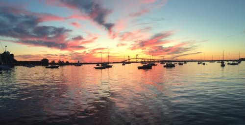 A serene sunset over a calm harbor, with boats silhouetted against vibrant pink and orange clouds.