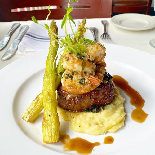 A plate featuring a filet mignon topped with shrimp, served with asparagus and creamy mashed potatoes, drizzled with sauce.