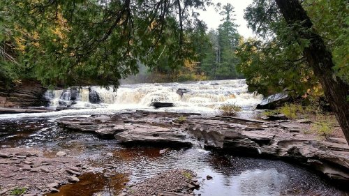 A serene waterfall cascades over rocky terrain, surrounded by lush greenery and autumn foliage.
