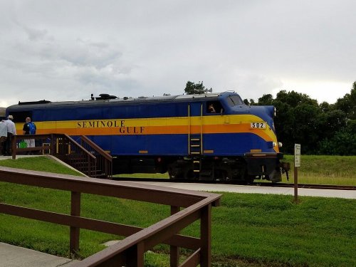 A blue and yellow Seminole Gulf train passes by a platform with people nearby on a cloudy day.