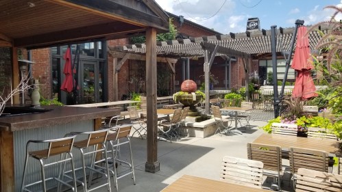 A sunny outdoor patio with wooden tables, chairs, and a fountain, surrounded by greenery and shaded by umbrellas.