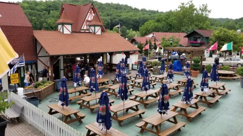 A spacious outdoor area with wooden picnic tables, surrounded by buildings and flags, featuring people in blue cloaks.