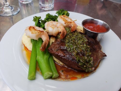 A plate featuring grilled shrimp, steak with herb sauce, broccoli, and celery, served with a side of sauce.