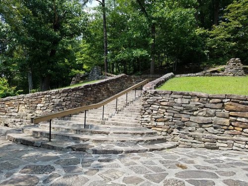 Stone steps with a railing lead up through a wooded area, surrounded by greenery and stone walls.