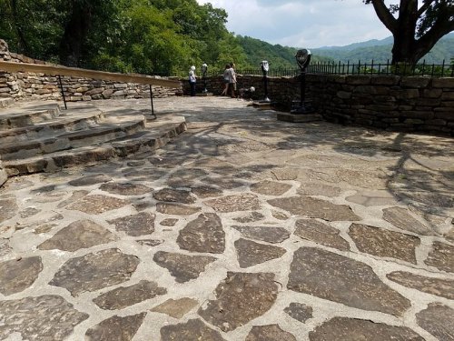 A stone pathway leads to a scenic overlook with trees and mountains in the background, featuring visitors enjoying the view.
