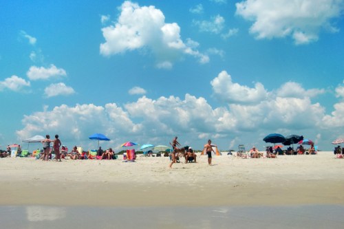 A sunny beach scene with people enjoying the sand, colorful umbrellas, and fluffy clouds in a blue sky.