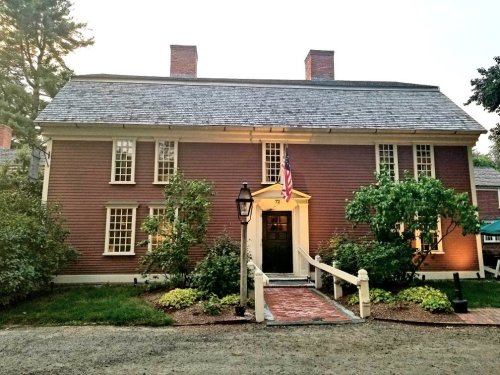 A historic red house with a slate roof, illuminated by lights, featuring a front porch and American flag.