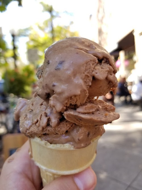 A hand holding a cone with two scoops of chocolate ice cream, set against a blurred outdoor background.