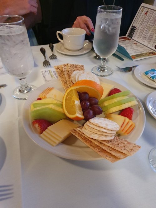 A colorful fruit platter with strawberries, grapes, orange slices, cheese, and crackers, accompanied by drinks and utensils.