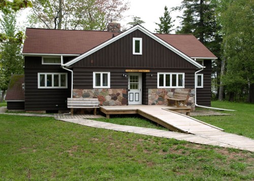 A brown wooden house with stone accents, surrounded by grass and trees, featuring a wooden walkway leading to the entrance.