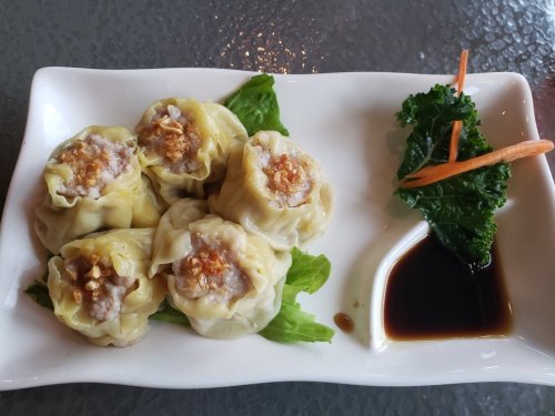 A plate of five steamed dumplings on lettuce, accompanied by a small dish of soy sauce and garnished with carrots and kale.