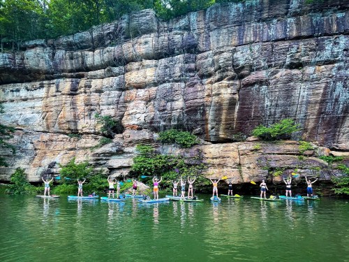 A group of people on paddleboards in a green lake, with a rocky cliff and trees in the background.