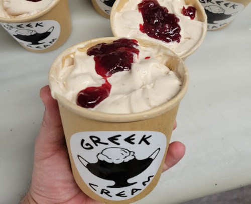 A hand holding a cup of Greek cream topped with red fruit jam, with more cups in the background.
