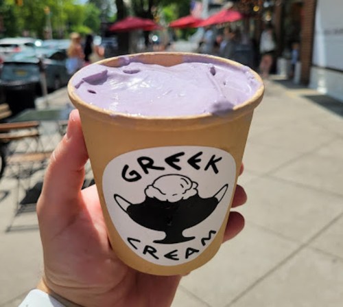 A hand holds a cup of purple ice cream labeled "Greek Cream" on a sunny street with people in the background.