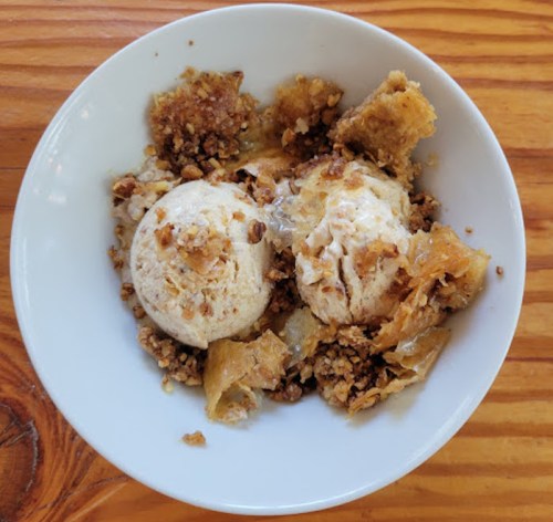 A bowl of dessert featuring two scoops of ice cream topped with crumbled pastry and nuts on a wooden table.