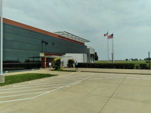 Modern building with large glass windows, two flags flying, and a paved area in front. Green fields in the background.