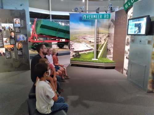 A family watches a video in a museum, seated in front of a display featuring a road sign and historical exhibits.