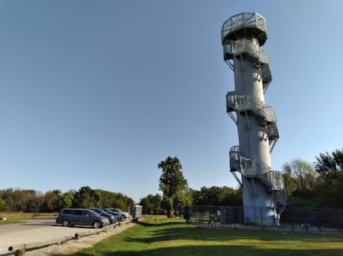A tall, gray observation tower with a spiral staircase beside a parking area and trees in the background.