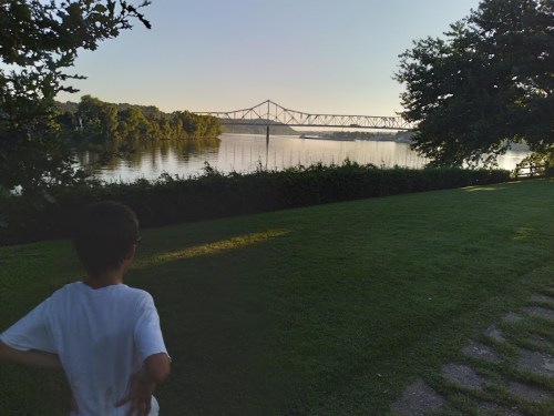 A child stands on a grassy path, looking at a river and a bridge in the distance during sunset.