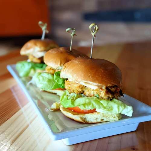 Three mini chicken sandwiches with lettuce and tomato, served on a white platter with decorative toothpicks.