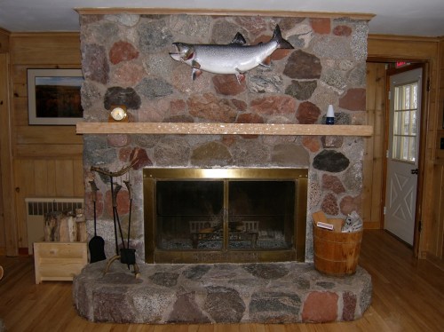 A rustic stone fireplace with a wooden mantel, featuring a mounted fish above and a cozy wooden interior.