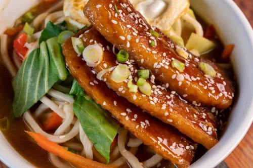 A bowl of noodles topped with glazed tofu, sesame seeds, green onions, and fresh vegetables in a savory sauce.