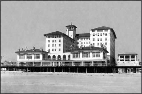 Historic black-and-white photo of a large seaside hotel with multiple stories and a raised wooden deck.