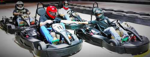 Four people in go-karts on a racetrack, wearing helmets and racing gear, reaching out to each other.