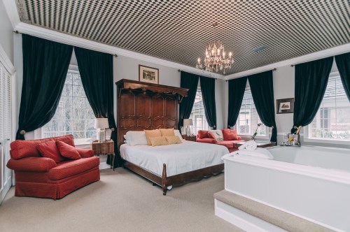 A spacious bedroom featuring a large bed, red armchairs, elegant curtains, and a chandelier.