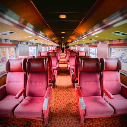 Interior of a vintage train car featuring rows of red upholstered seats and patterned carpet.