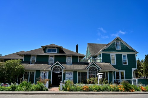 A charming green house with a white picket fence, featuring a large porch and colorful flowers in the front yard.